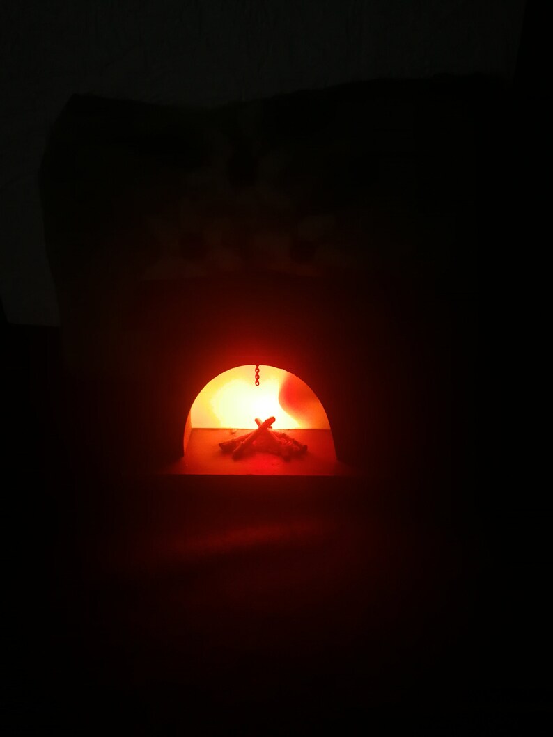 May include: A miniature fireplace with a glowing orange interior. The arched opening reveals a small pile of wooden logs illuminated by a warm light. A chain hangs from the top of the opening. The background is dark.