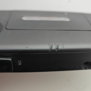 May include: Black cassette player with a silver button labelled "REC" on the side. The player has some scratches on the side.