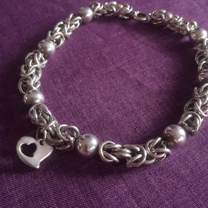 May include: A silver bracelet with a heart charm. The bracelet is made of a chain with small round beads. The heart charm is open and has a delicate design.