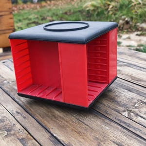 Könnte beinhalten: Ein roter und schwarzer Kunststoff-Aufbewahrungsbehälter mit mehreren Fächern. Der Behälter ist quadratisch mit schwarzer Oberseite und roten Trennwänden. Er steht auf einer verwitterten Holzoberfläche.