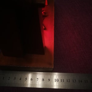 May include: A wooden object with a red glow, possibly a lamp or decorative piece. A metal ruler with inch markings is placed near the object, showing measurements up to 6 inches. The object is on a dark purple surface.