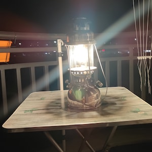 Könnte beinhalten: Eine schwarze Laterne mit einem Glasschirm steht auf einem Holztisch mit einem Blumenmuster. Die Laterne ist angezündet und strahlt ein warmes Licht aus.