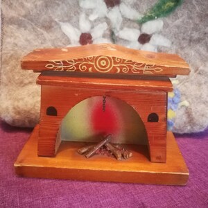 May include: A miniature wooden fireplace with a decorative design on the top. The fireplace has a red and green painted interior with small wooden logs inside. The base is rectangular and the overall color is brown.