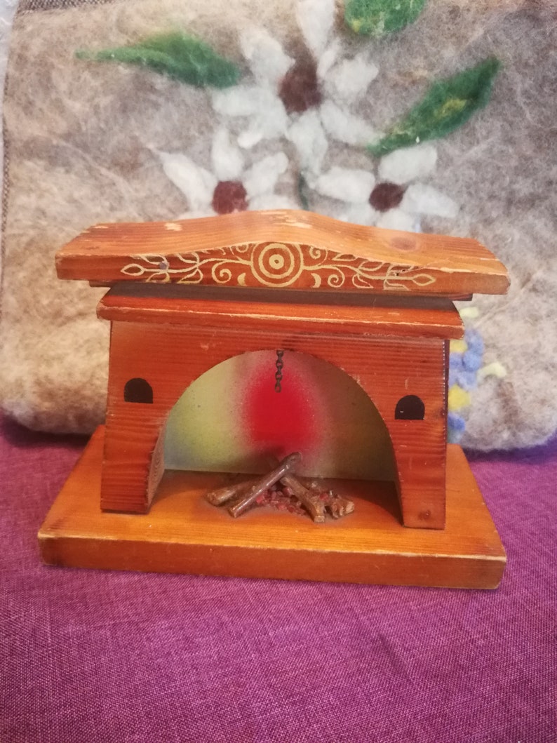 May include: A miniature wooden fireplace with a decorative design on the top. The fireplace has a red and green painted interior with small wooden logs inside. The base is rectangular and the overall color is brown.