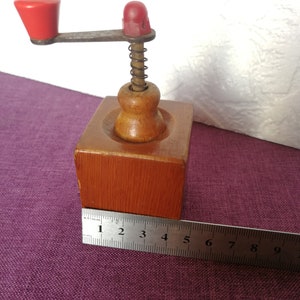 May include: A small wooden coffee grinder with a red crank handle and a spring mechanism. The grinder is about 5 inches tall and is made of natural wood.