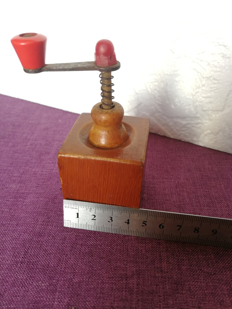May include: A small wooden coffee grinder with a red crank handle and a spring mechanism. The grinder is about 5 inches tall and is made of natural wood.