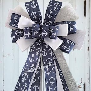May include: A decorative bow featuring navy blue ribbon printed with white anchors, white satin ribbon, and natural burlap ribbon. The bow is designed with multiple loops and tails, perfect for nautical-themed decor. The text "Emerald Waters Wreath Decor" is visible on the ribbon.