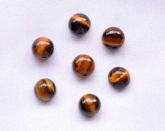 May include: Seven polished, round tiger's eye gemstones. The stones display a mix of brown, gold, and black colors, with a glossy finish. The gemstones are arranged on a white, textured surface, showcasing their natural patterns and colors.