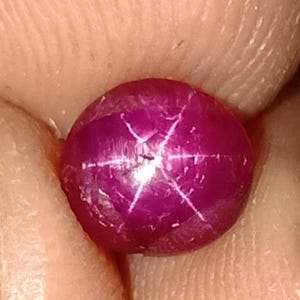 以下が含まれることがあります： 6条の星模様が特徴的な、丸く濃いマゼンタ色の宝石。指の間に挟まれ、研磨された表面と光の反射によって生み出される星の効果が示されています。宝石の色は豊かで鮮やかな色合いです。