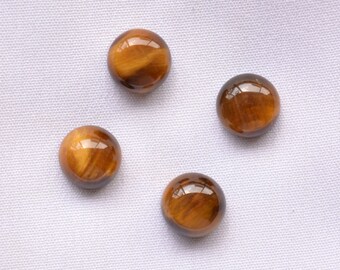 May include: Four polished, round tiger's eye gemstones. The stones are a warm brown color with golden and black streaks, and have a glossy, reflective surface. They are arranged on a white fabric background.