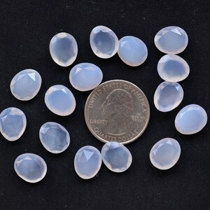 Puede incluir: Un conjunto de gemas ovaladas de color azul p&aacute;lido con superficies facetadas, dispuestas sobre un fondo negro. Se incluye una moneda para comparar el tama&ntilde;o. Las gemas tienen una cualidad transl&uacute;cida.
