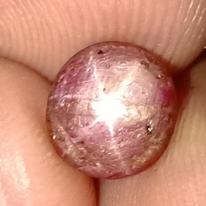 May include: A close-up of a cabochon-cut, star ruby gemstone. The gem is a mix of pink and purple hues, with a prominent six-rayed star effect visible across its surface. The stone is held between fingers.