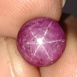 May include: A round, polished, purplish-red gemstone with a six-rayed star effect. The star pattern is created by light reflecting off inclusions within the stone. The gem is held between fingers, showcasing its color and clarity.