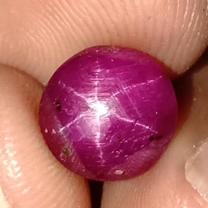Puede incluir: Una gema pulida, redonda, de color magenta intenso, con un efecto de estrella de seis rayos. La gema se sostiene entre los dedos, mostrando su color vibrante y su fenómeno óptico único. El patrón de estrella es resultado de la reflexión de la luz.