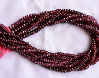 May include: Strands of faceted, deep red garnet beads. The beads are small and round, strung together to form multiple strands. A pink tassel is attached to the end of the strands. The beads have a polished, reflective surface.