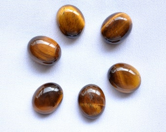 Könnte beinhalten: Sechs polierte, ovale Tigeraugen-Edelsteine, kreisförmig angeordnet. Die Steine zeigen eine satte, goldbraune Farbe mit Chatoyanz, die das Licht wie ein Katzenauge reflektieren. Die Edelsteine liegen auf einem weißen Hintergrund.
