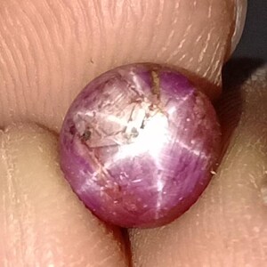 May include: A close-up of a round, polished gemstone with a star effect. The gem is a deep purplish-red colour with a six-rayed star pattern radiating from the centre. The surface has some natural inclusions and imperfections.