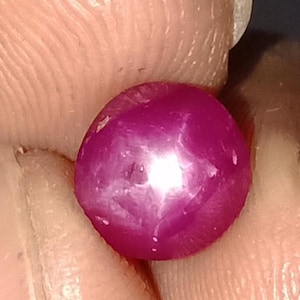 May include: A close-up of a round, polished, magenta-colored gemstone. The gem has a star-like effect in the center, with a bright white highlight. The gem is held between two fingers.