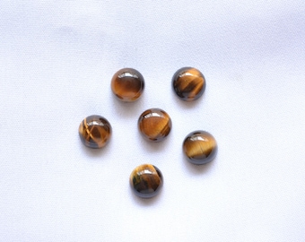May include: A collection of six polished tiger's eye gemstones, each with a rounded shape. The stones display a mix of brown and gold hues, with visible chatoyancy. They are arranged on a white, textured surface.