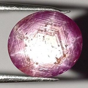 May include: A round, polished ruby gemstone held by silver tweezers. The ruby is a deep pinkish-red color with a star-like pattern visible on its surface. The gemstone is smooth and has a slight sheen.