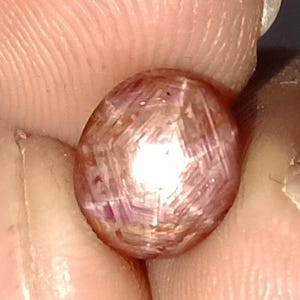 May include: A close-up of a pink gemstone held between fingers. The gemstone has a rounded shape with a star-like pattern visible on its surface. The gem's color is a mix of pink and light brown.