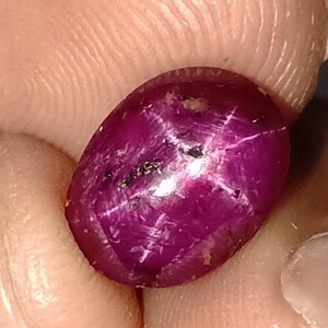 Puede incluir: Una gema de rubí pulida, de forma ovalada. La gema es de un color magenta profundo y saturado, con un efecto de estrella de seis rayos. La piedra se sostiene entre los dedos, mostrando su belleza natural y su patrón estelar único.