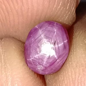May include: A close-up of a polished, oval-shaped, purplish-pink gemstone. The gem has a star-like effect, with lines radiating from a central point. The surface is smooth and reflective, with a slight translucence.