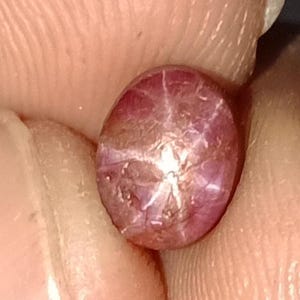May include: Oval-shaped, reddish-purple star ruby gemstone. The gem displays a six-rayed star pattern across its surface. The stone is held between fingers, showcasing its polished, reflective qualities. A close-up view highlights the gem's natural beauty.