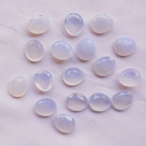 Puede incluir: Una colecci&oacute;n de gemas ovaladas de color blanco azulado p&aacute;lido con superficies facetadas. Las gemas est&aacute;n dispersas sobre una tela blanca texturizada, mostrando su sutil color y corte. La disposici&oacute;n resalta las piedras individuales.