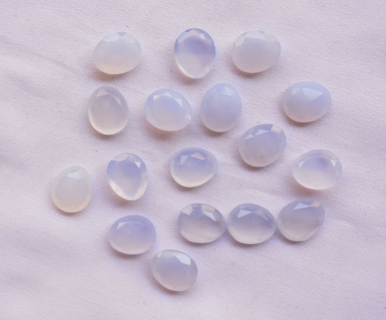 Puede incluir: Una colecci&oacute;n de gemas ovaladas de color blanco azulado p&aacute;lido con superficies facetadas. Las gemas est&aacute;n dispersas sobre una tela blanca texturizada, mostrando su sutil color y corte. La disposici&oacute;n resalta las piedras individuales.