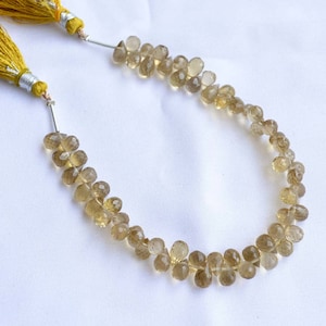 May include: A necklace of faceted, teardrop-shaped beads in shades of amber and brown. The beads are strung together, creating a delicate, curved shape. The necklace is finished with gold-colored tassels on each end.