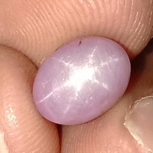 May include: Oval-shaped, light purple gemstone with a six-rayed star pattern. The gem is smooth and polished, reflecting light across its surface. The star effect is prominent, creating a captivating visual display. This gemstone is likely a star sapphire or ruby.