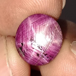 Könnte beinhalten: Nahaufnahme eines polierten, oval geformten Edelsteins. Der Edelstein hat eine tiefmagentafarbene Farbe mit einer hellen, reflektierenden Oberfläche. Feine, lineare Einschlüsse erzeugen einen sternartigen Effekt. Der Edelstein wird zwischen zwei Fingern gehalten.