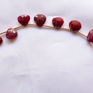 May include: Strung ruby gemstone beads. The faceted, heart-shaped beads are a deep red color and are strung on a gold-colored wire. The beads are displayed on a white, slightly textured surface.