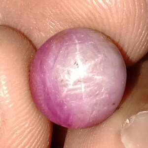 May include: A close-up of a round, polished gemstone with a star effect. The gem is a mix of pink and white hues, with a six-rayed star pattern visible on its surface. The gem is held between fingers.