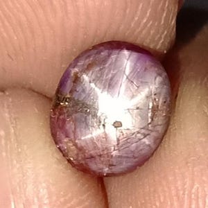 Puede incluir: Una gema pulida de forma ovalada con efecto estrella. La gema tiene un tono rojo púrpura con un patrón de estrella blanco brillante en su superficie. Se ven líneas finas e inclusiones dentro de la piedra, lo que aumenta su belleza natural.