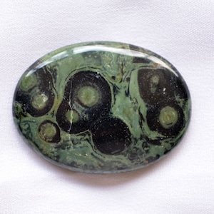 Könnte beinhalten: Oval geformter Edelstein mit polierter Oberfläche. Der Stein weist ein Muster aus dunkelgrünen und schwarzen kreisförmigen Designs auf, die Augen oder Flecken ähneln, vor einem helleren grünen Hintergrund. Der Stein ist etwa 7,6 cm lang.