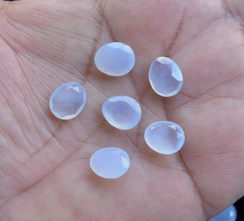 Puede incluir: Seis gemas ovaladas de color azul p&aacute;lido con superficies facetadas se muestran en una mano. Las gemas tienen una calidad transl&uacute;cida, reflejando un tono suave y fresco. Ideal para la creaci&oacute;n de joyas.
