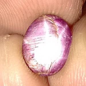 May include: Oval-shaped, polished gemstone with a vibrant pink hue. The gem has a smooth, reflective surface with internal striations and a bright, white area. The gemstone is held between two fingers.