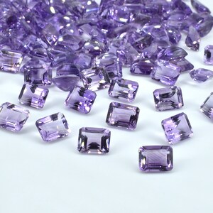 May include: A collection of rectangular, faceted amethyst gemstones in varying sizes. The gems are a translucent purple hue, scattered across a white surface. The light catches the facets, creating highlights and shadows.