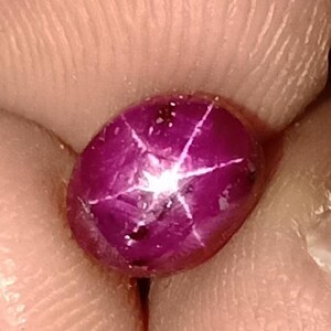 Może przedstawiać: Zbliżenie wypolerowanego, owalnego, głębokiego, purpurowego kamienia szlachetnego. Klejnot ma efekt sześcioramiennej gwiazdy z jasnymi białymi liniami promieniującymi z centralnego punktu. Klejnot jest trzymany między palcami.