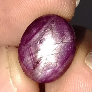May include: A polished, oval-shaped ruby gemstone. The gem is a deep purplish-red color with a bright, reflective surface. Visible internal lines and inclusions add to its unique character. The stone is held between fingers.