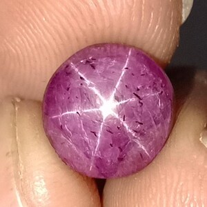 May include: A round, polished, star ruby gemstone. The gem is a deep pinkish-purple color with a six-rayed star pattern radiating from the center. The star is white and the gemstone is held between fingers.