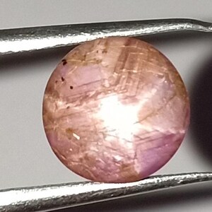 May include: A round, polished gemstone held by tweezers. The gem has a pinkish-brown hue with internal patterns and inclusions. The surface reflects light, creating a subtle shimmer. The tweezers are silver-colored.