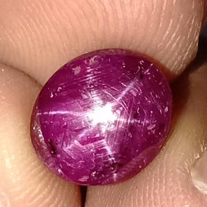 Puede incluir: Primer plano de una gema de color magenta oscuro con un efecto de estrella. La piedra de forma ovalada tiene una superficie pulida con inclusiones visibles y un patrón de estrella de seis rayos. La gema se sostiene entre los dedos, resaltando su color y forma.
