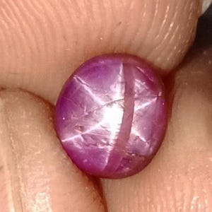 May include: A close-up of a polished, oval-shaped, purplish-red gemstone. The gem features a six-rayed star effect, with a bright, white star pattern visible across its surface. The stone is held between fingers.