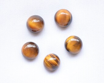 May include: Five polished, round tiger's eye gemstones. The stones are a warm brown color with golden-yellow bands and a glossy finish. The gemstones are arranged on a white surface.