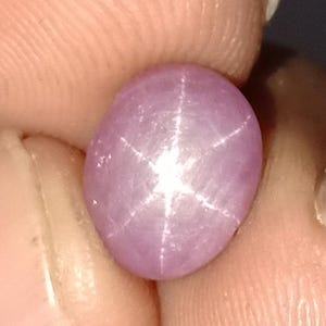 以下が含まれることがあります： 楕円形、紫がかったピンク色の宝石で、6条の星模様があります。星の効果は、石内のインクルージョンからの光の反射によって生まれます。磨かれた表面と独特の光学現象を示すために、宝石は指の間に保持されています。