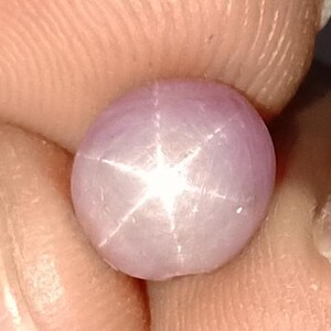 May include: A round, light purple gemstone with a six-pointed star pattern. The star is white and radiates from the center of the stone. The gem is held between fingers, showing the texture of the skin.