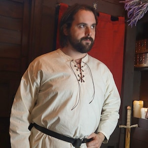 May include: A white long-sleeved shirt with a lace-up neckline and a brown leather belt. The shirt is being worn by a person with a beard and is standing in front of a wooden wall.